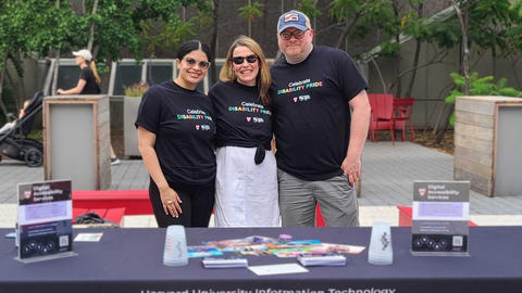 Suany, Janell, and Derek stand and smile behind the DAS Table on the Science Center Plaza. All are wearing "Celebrate Disability Pride" t-shirts.