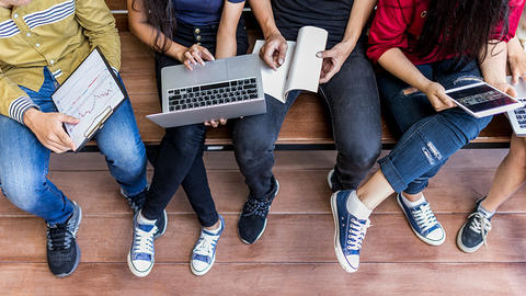 Students sitting on a bench using high and low-tech devices including a laptop, tablets, a notebook, and paper on a clipboard.