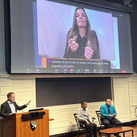 An ASL interpreter and captions are on a large projection screen above Michael speaking from a lectern and Jennison and Kyle seated nearby.