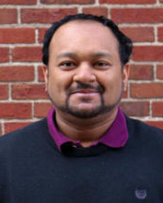 Caesar, a medium olive‑skinned man with a trimmed beard and short hair, wears a black sweater over a purple shirt and stands in front of a red brick wall, smiling softly.