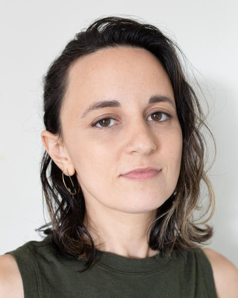 Kay, with dark wavy hair and light olive skin, in front of a soft white background. They are smiling subtly, wearing thin gold hoop earrings and a sleeveless olive green top.