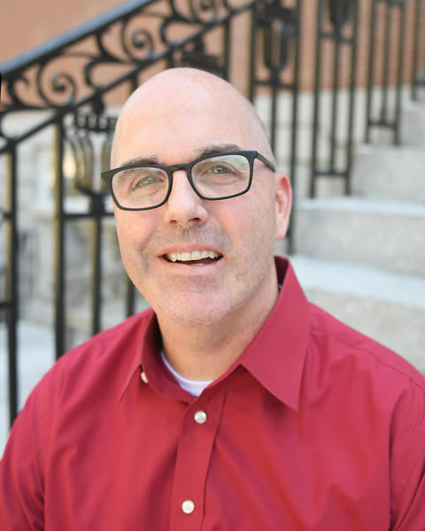 Erich smiles for the camera. He is a middle-aged, white man with a shaved head and dark rimmed glasses, wearing a button down crimson with a collar.