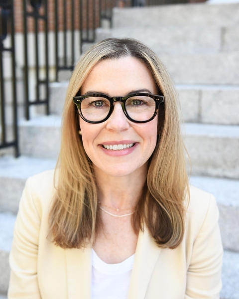 A smiling woman with fair skin and shoulder-length blonde hair and dark-rimmed glasses is seated on stone steps. She is wearing a light-colored blazer over a white top, giving her a professional and approachable appearance.