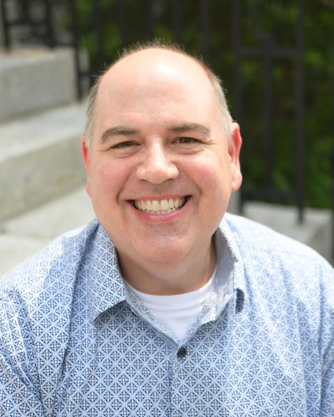 Curtis Wilcox, a smiling bald man wearing a blue patterned shirt sitting outdoors on stone steps.