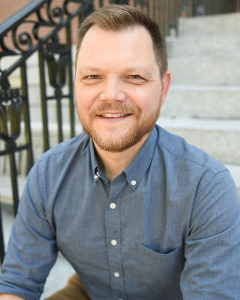 A man seated on outdoor stone steps, smiling warmly at the camera. He has light skin, short light-brown hair, and a neatly trimmed beard. He is wearing a blue-gray button-down shirt with the sleeves rolled up.