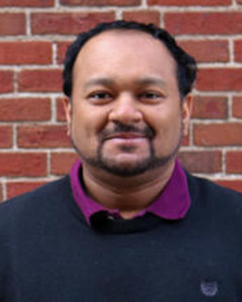 Caesar, a medium olive‑skinned man with a trimmed beard and short hair, wears a black sweater over a purple shirt and stands in front of a red brick wall, smiling softly.