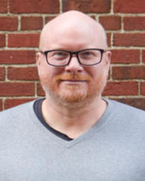 Derek's headshot shows him in front of a brick wall. He is a middle aged, white man with a shaved head and dark framed glasses. He has some reddish stubble that highlights his engaging smile. 
