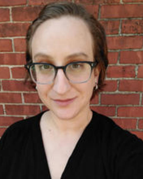 Close cropped headshot of woman with dark brown hair and glasses, against brick wall. 
